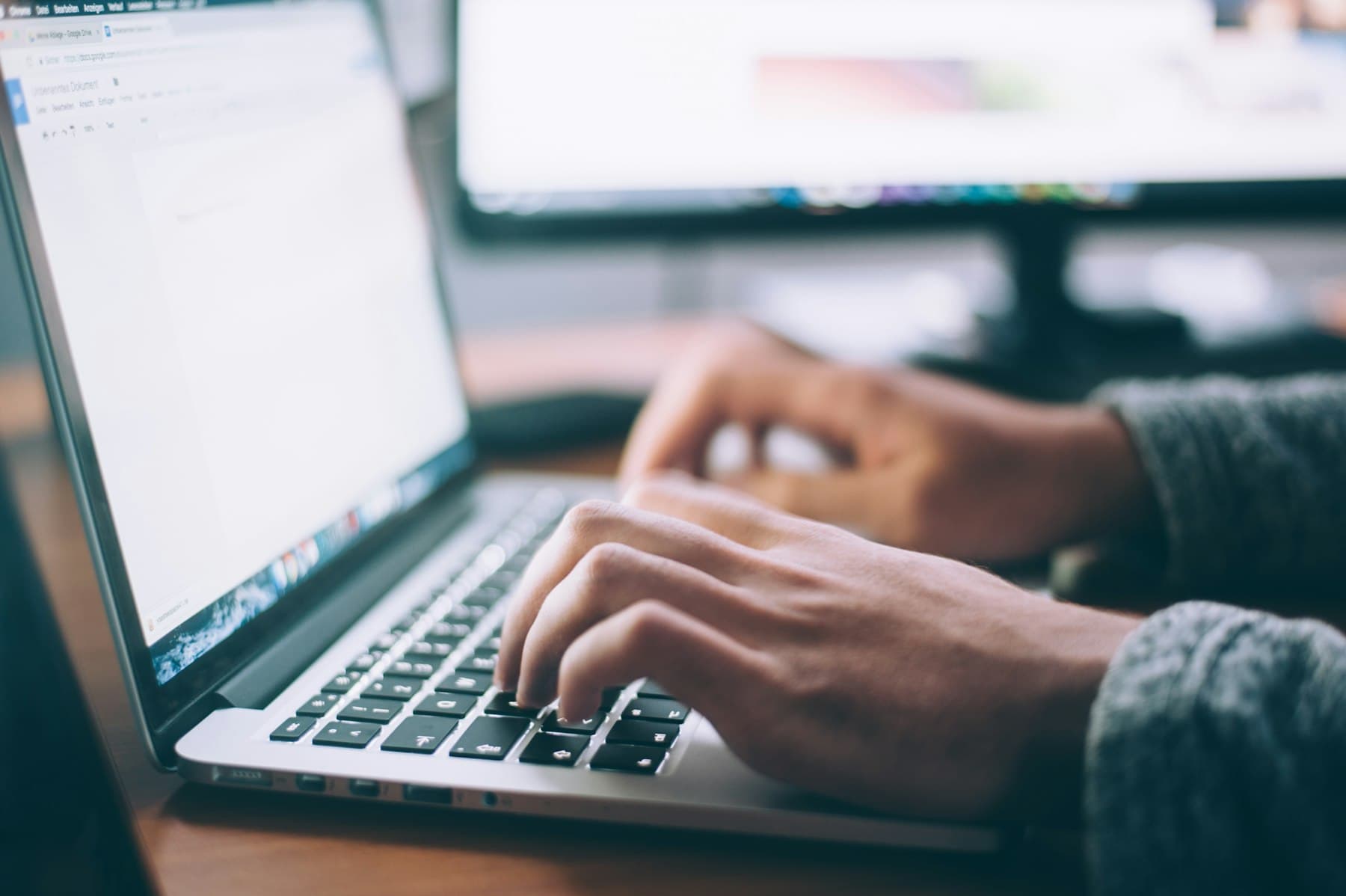 Close-up of hands typing on a laptop keyboard, with a document visible on a blurred screen in the background