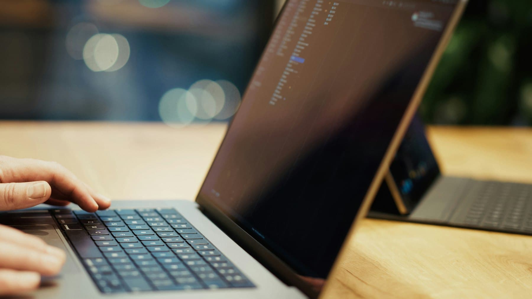 Close-up of a hand typing on a laptop keyboard on a wooden table, with a houseplant blurred in the background