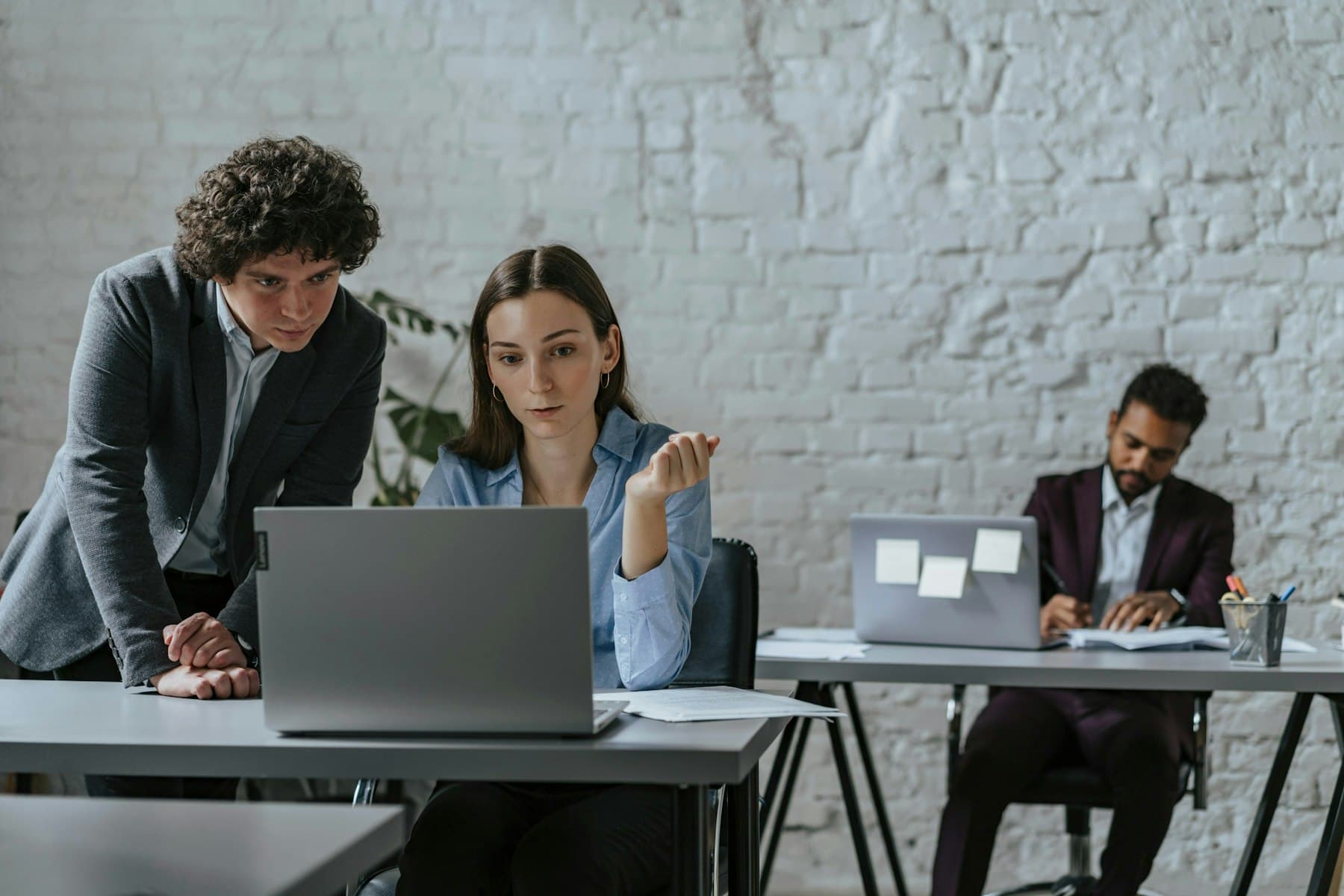 A colleague leans in beside a coworker who is looking at a laptop screen together with him while explaining something