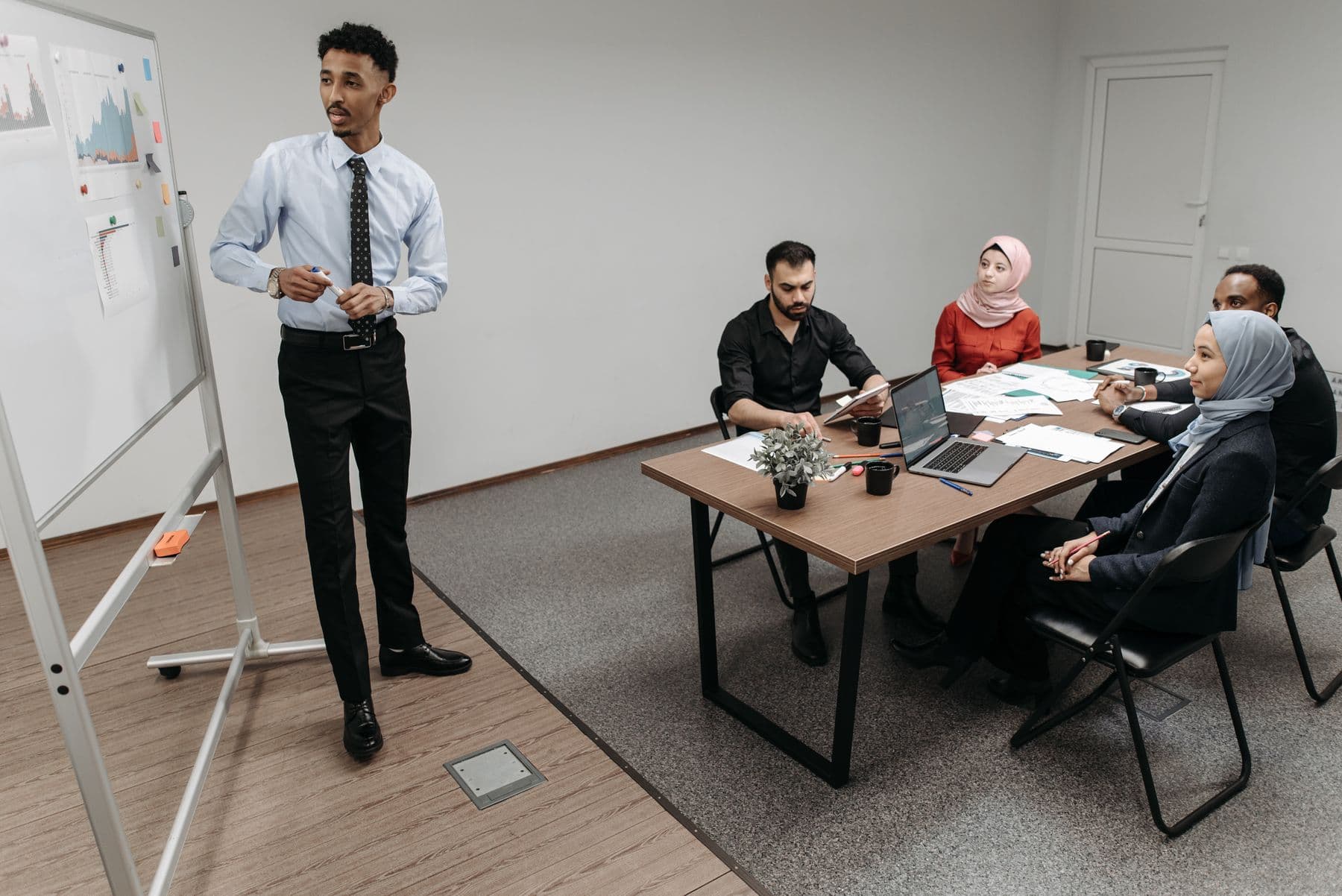 Trainer explains charts on a flipchart to a small, diverse group of office employees, with a meeting table and laptop in the background