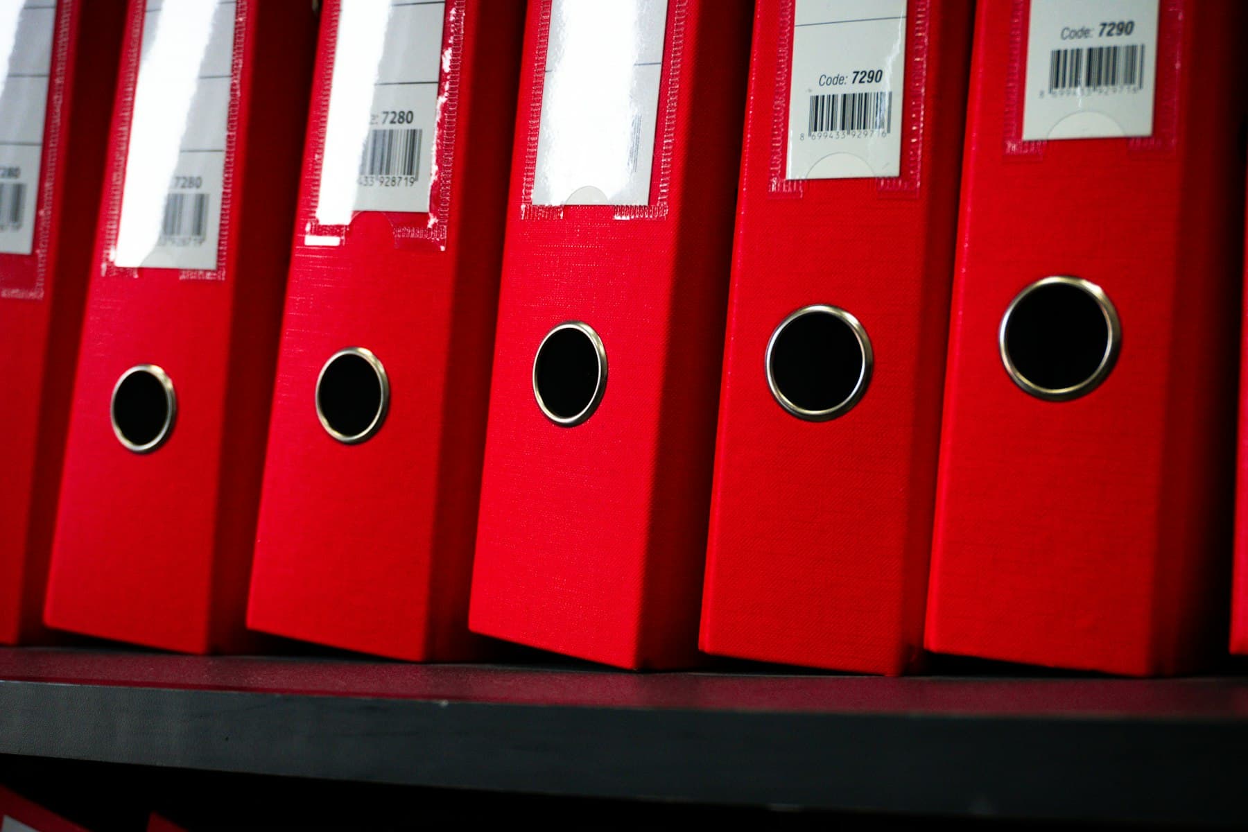 A row of neatly arranged red ring binders with label tags on a shelf