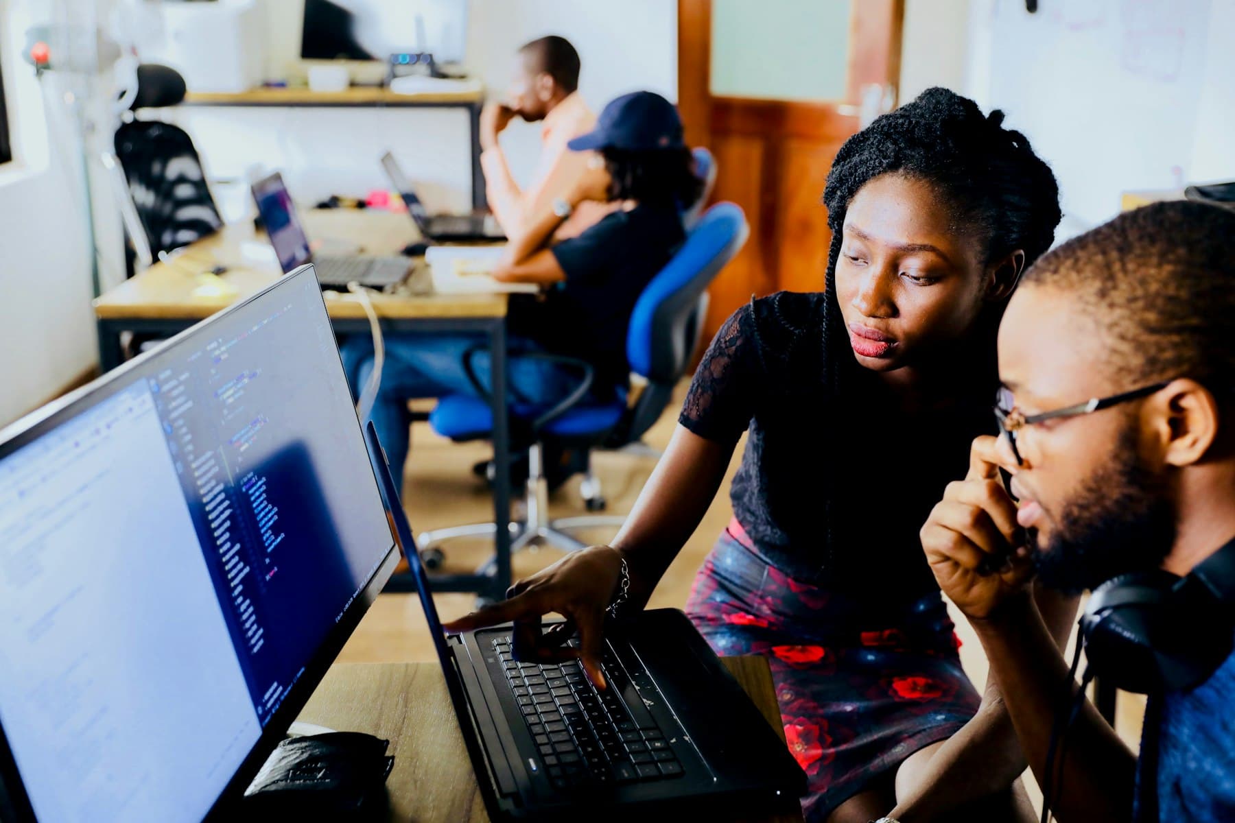 A woman and a man look together at a monitor showing source code, she operates the laptop touchpad